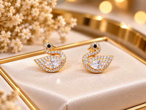 A pair of gold-toned swan-shaped stud earrings with clear pavé stones and small black eye accents, displayed on a cream satin jewelry tray with decorative flowers and warm blurred lights in the background.